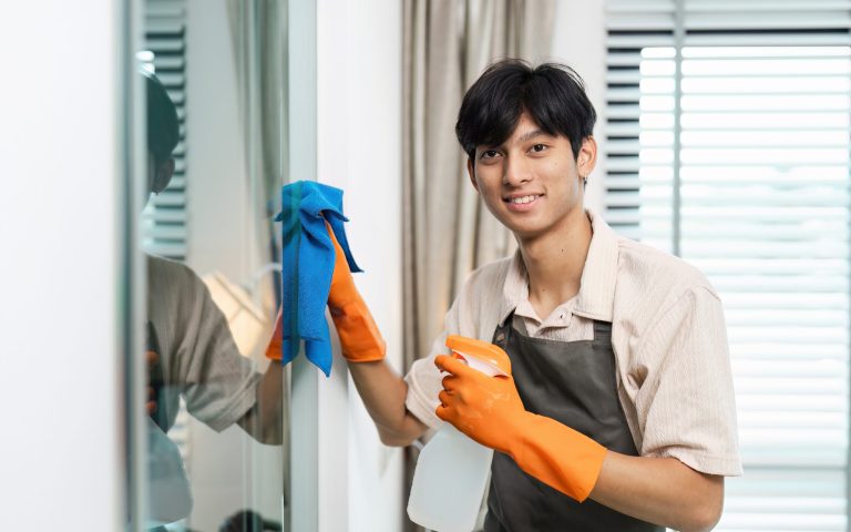 A young man in an apron and gloves cleans a window with a spray bottle and cloth, smiling at the camera in a modern home interior.